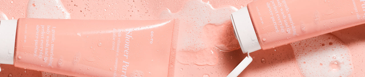 Two pink cosmetic tubes with white caps lie on a wet, bubbly pink surface, partially covered in foam and water droplets. The tubes are angled and have text printed on them.