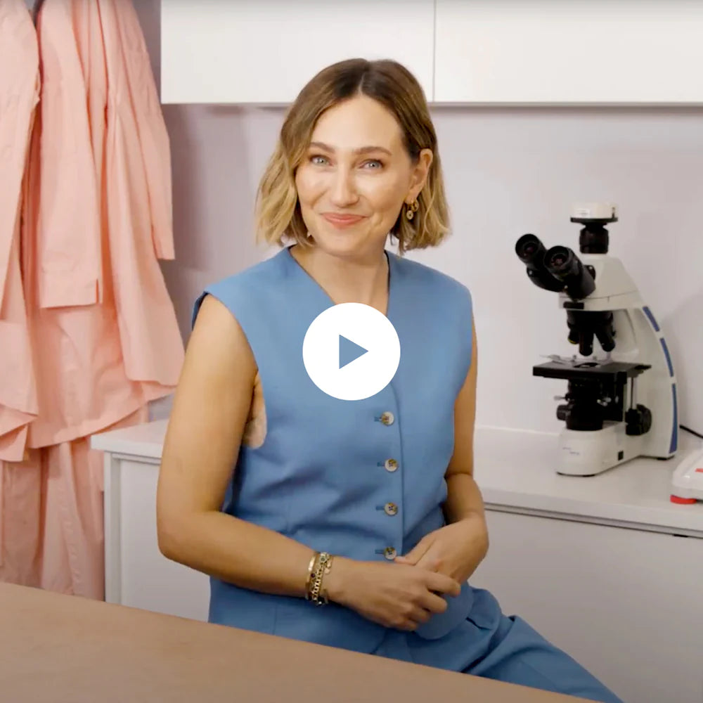 A woman in a blue sleeveless top sits beside a microscope in a lab, smiling at the camera. Pink lab coats hang in the background. A play button icon is centered on the image.