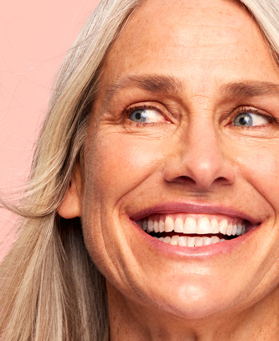 A close-up of a smiling older woman with long gray hair and blue eyes, showing her teeth. She looks off to the side against a soft pink background.