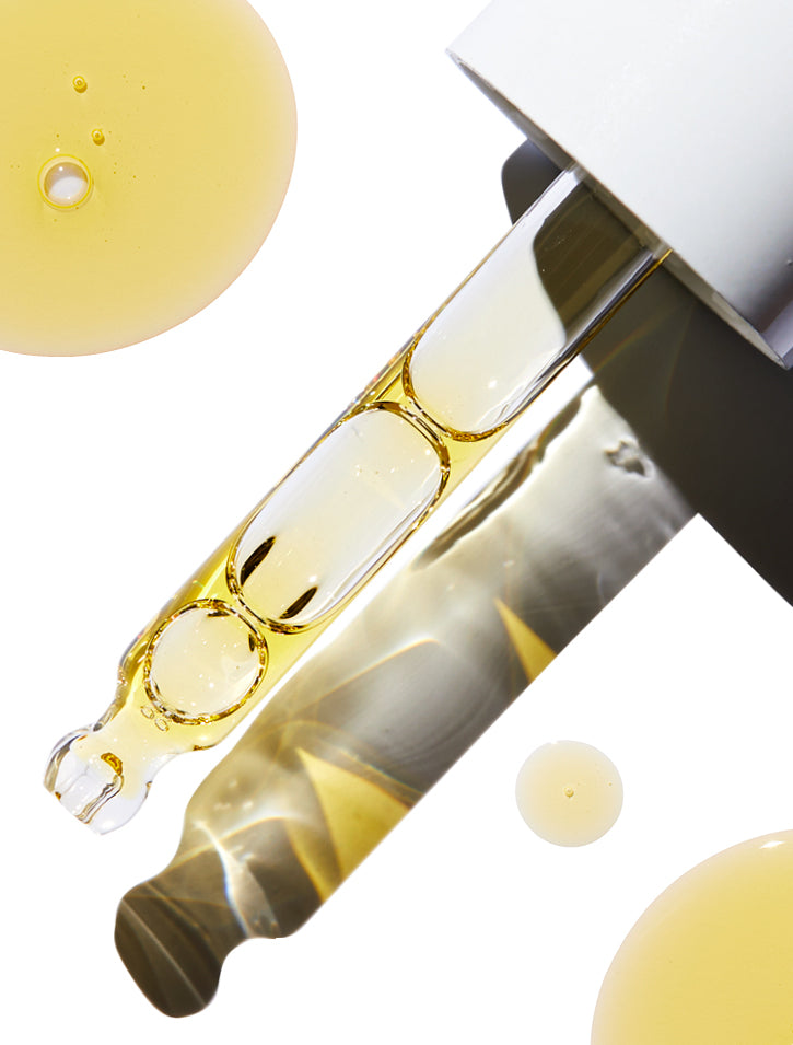 A close-up of a dropper filled with golden liquid, surrounded by drops of the same liquid on a white background. The dropper has a shiny metallic cap.