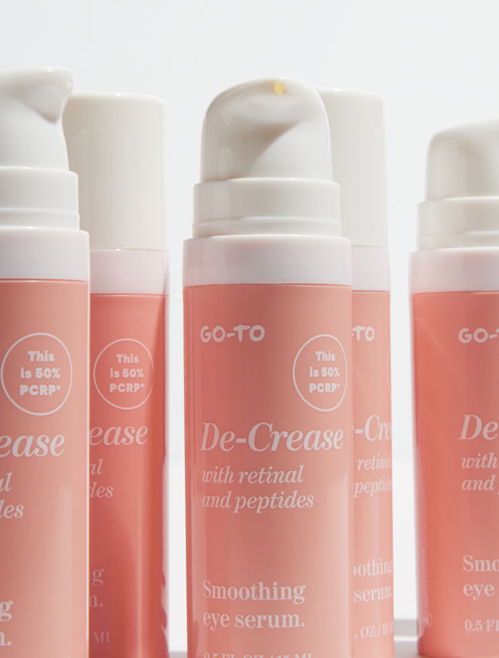 Four bottles of Go-To De-Crease Smoothing Eye Serum with retinal and peptides, featuring pink and white packaging and labels indicating the use of 50% PCR (post-consumer recycled) materials.