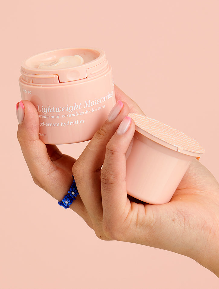 A hand holds an open jar of lightweight moisturizer with a light pink container, while the other hand holds the lid. The background is a matching soft pink. A blue beaded ring is visible on one finger.