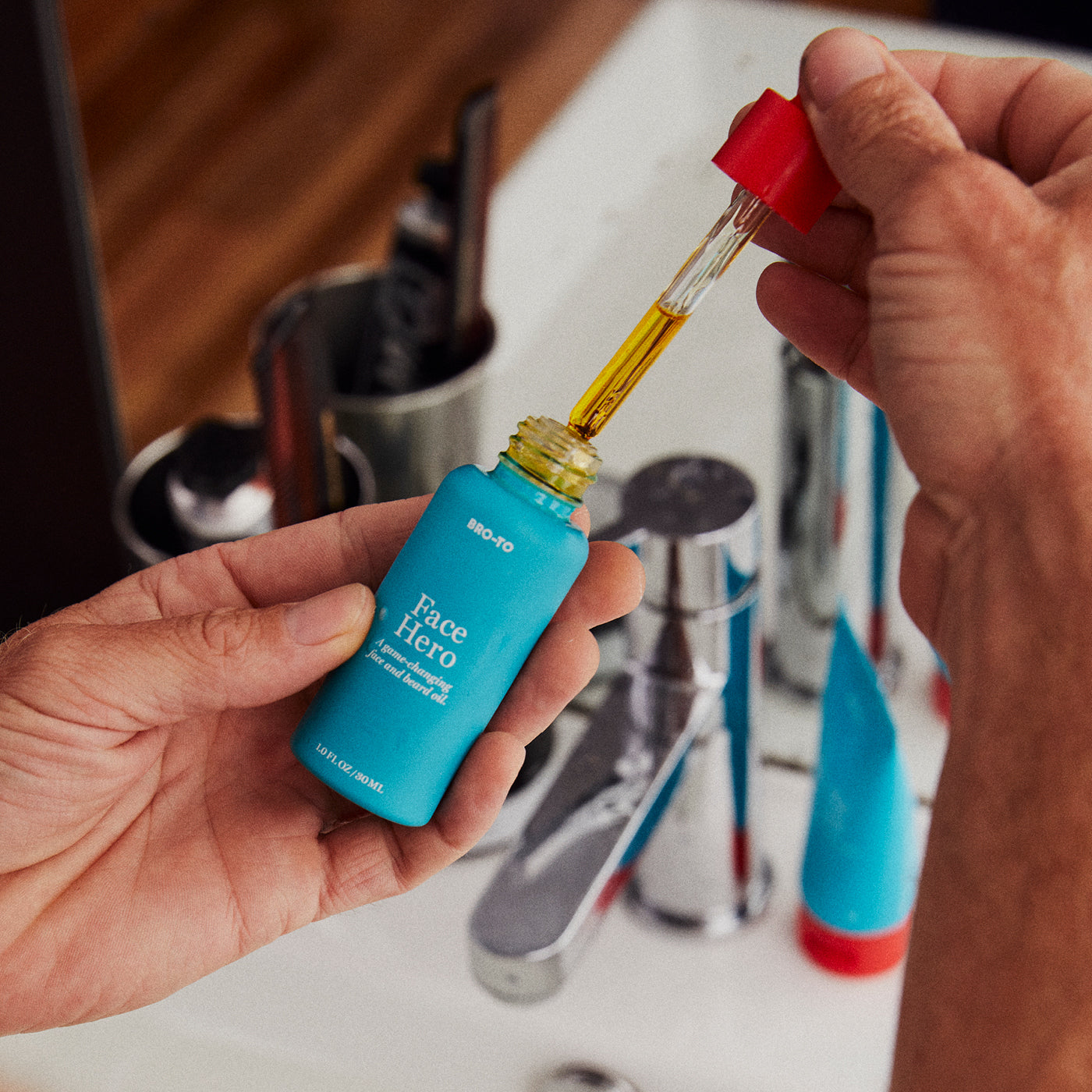 A person holds a blue bottle labeled Face Hero and uses a dropper to extract yellow oil, with a sink and bathroom items visible in the background.