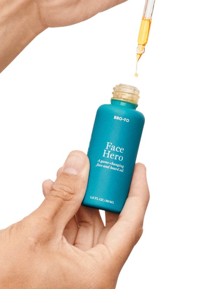A hand holds a blue bottle labeled Face Hero while another hand uses a dropper to dispense golden facial oil from the bottle against a white background.