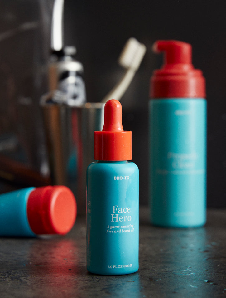 A blue bottle labeled Face Hero with a red dropper cap stands on a counter. Two more blue and red skincare bottles are blurred in the background. The setting appears to be a bathroom.