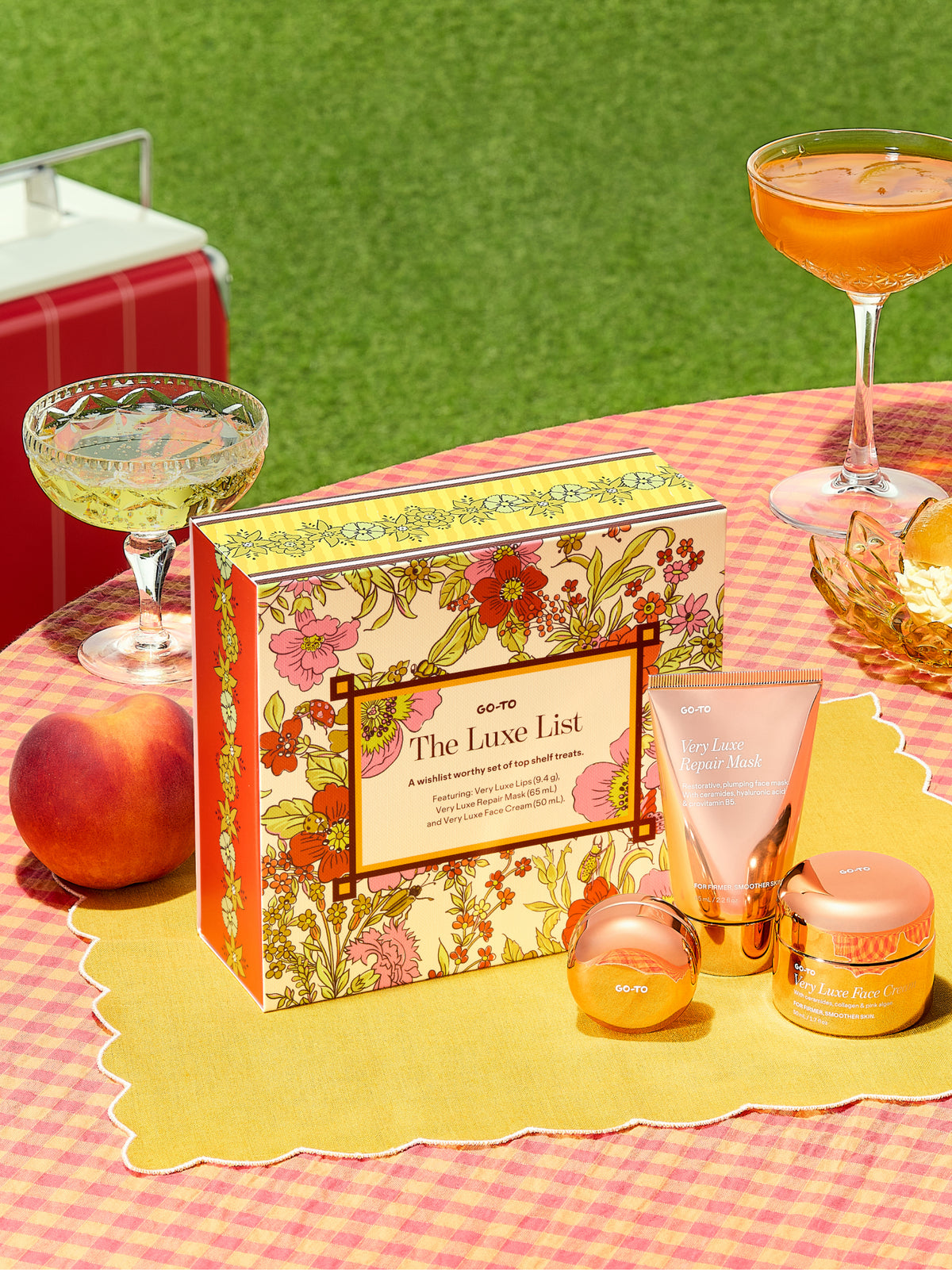Go-To Skincare The Luxe List floral gift set with face cream lip balm and repair mask displayed on a yellow placemat beside colorful drinks.