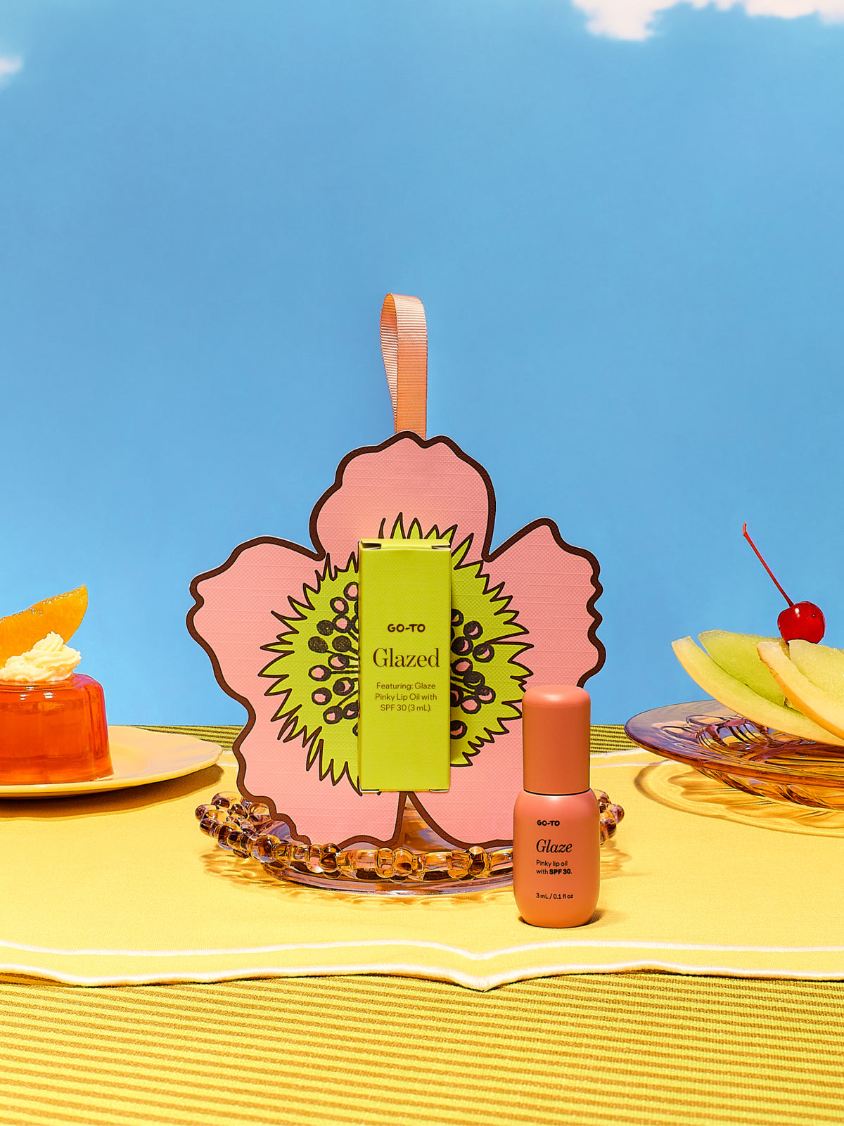 Go To Skincare Glazed tinted SPF lip oil and gift packaging displayed with a pink flower card orange tube citrus fruit and cherry on a sunny yellow table