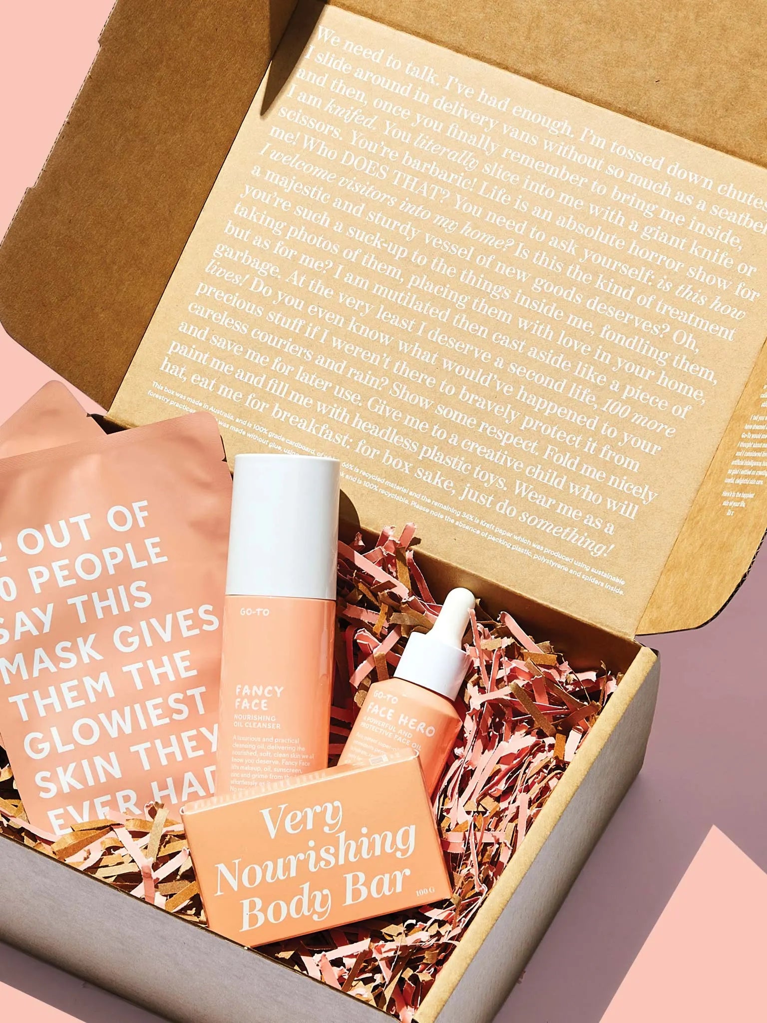 An open cardboard box with shredded paper contains skincare products in peach-colored packaging, including a face mask, serum, and body bar. The inside lid features printed text, and the box sits on a pale pink surface.