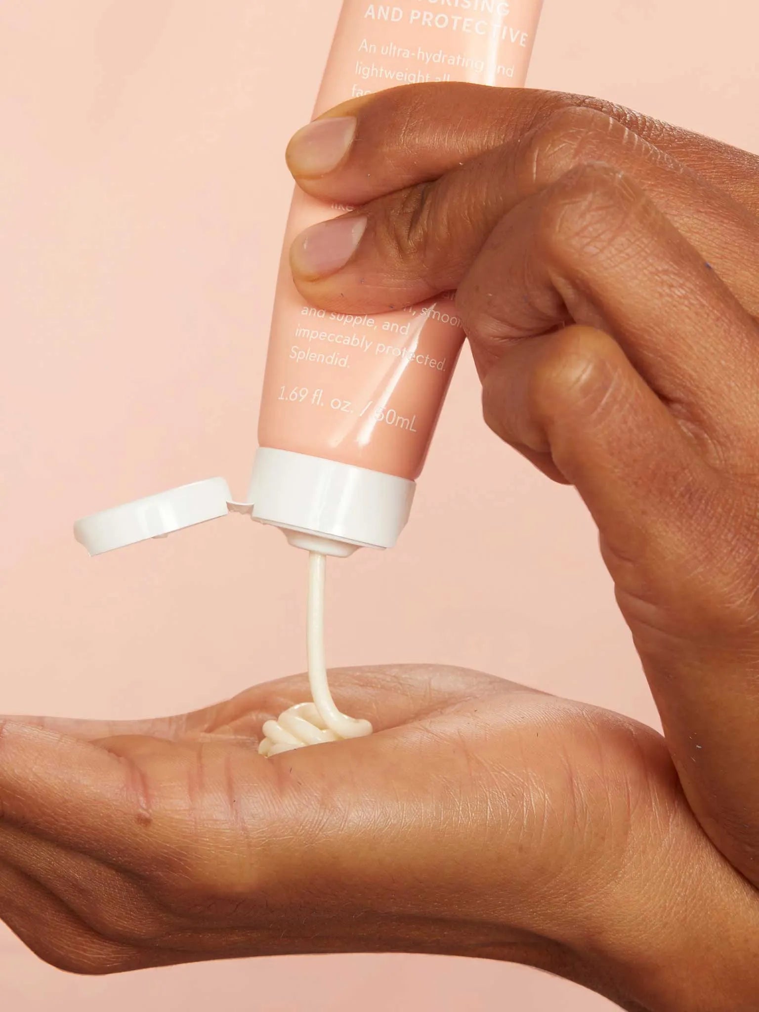 A person squeezes a creamy lotion from a peach-colored tube onto their palm against a light pink background.