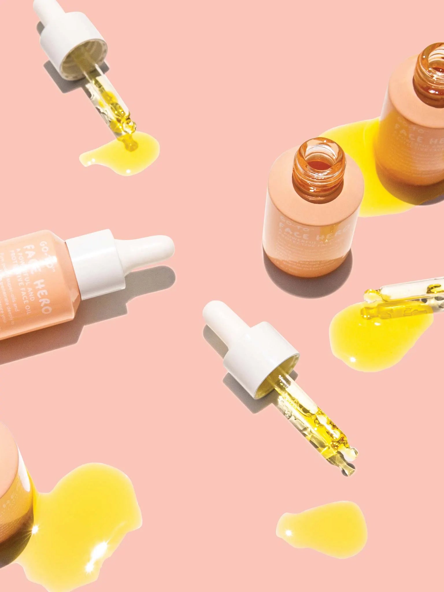 Three dropper bottles of yellow face oil are open and tipped over on a peach background, with droppers and spilled oil scattered around them.
