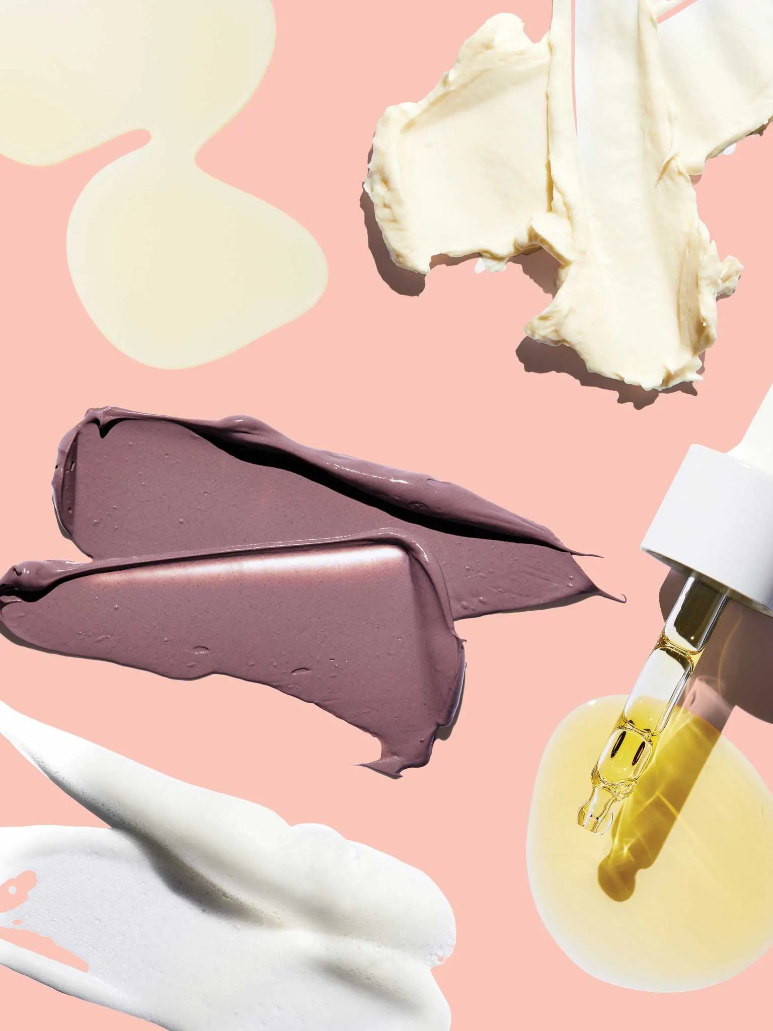 Various skincare creams, lotions, and a yellow serum with a dropper are artfully smeared and dripped on a pale pink background in an abstract arrangement.