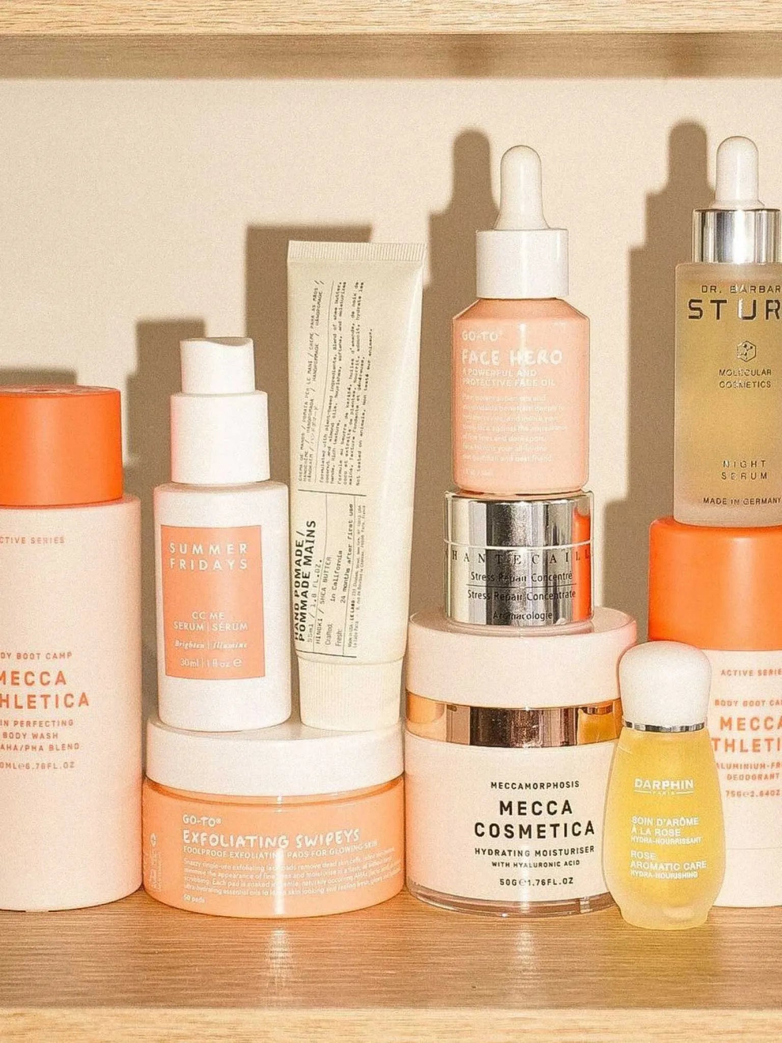 A shelf displaying various skincare products in pastel and neutral containers, including bottles, jars, and tubes labeled with brands such as Mecca Cosmetica, Summer Fridays, and Go-To.
