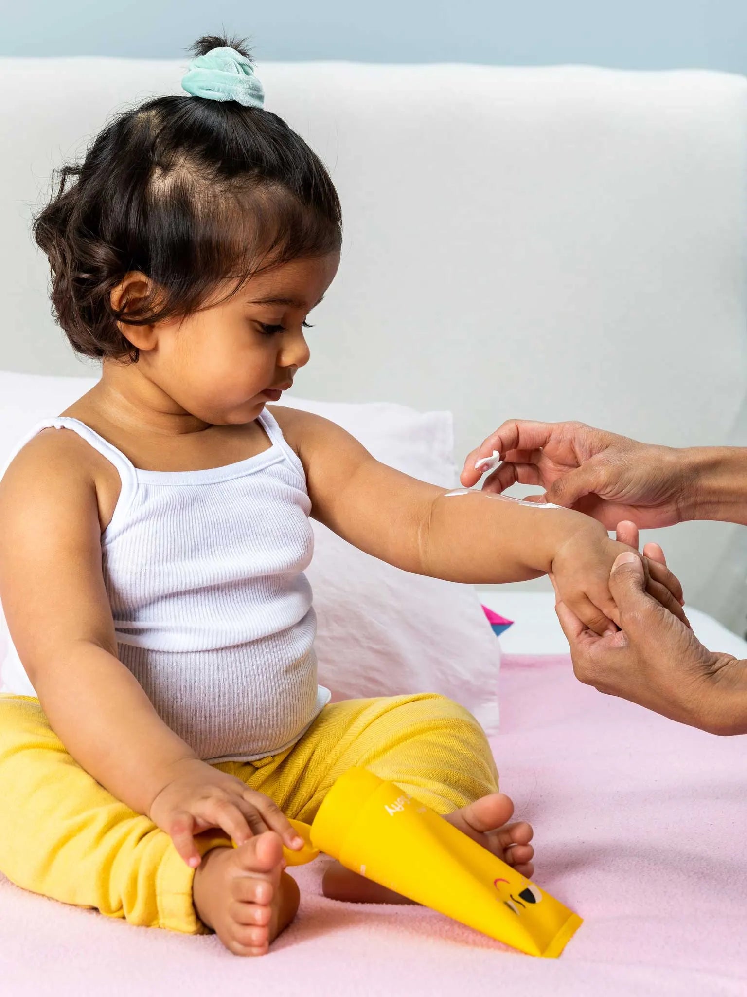 A young child sits on a bed in yellow pants and a white tank top while an adult applies lotion to the child’s arm. A yellow lotion tube rests in the child’s lap.