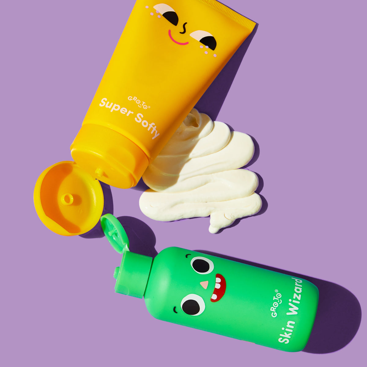 Two colorful bottles of skincare products with smiling cartoon faces lie on a purple background. The yellow “Super Softy” tube dispenses white cream, while the green “Skin Wizard” bottle is closed.