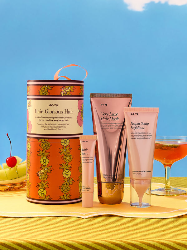 Go-To Skincare Hair Glorious Hair gift set with an orange patterned box and three pink hair care tubes displayed on a sunny table beside fruit and a cocktail