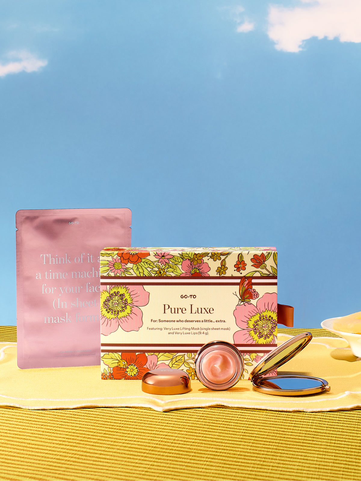 Go To Skincare Pure Luxe gift set with floral packaging a pink mask pouch and skincare products displayed on a sunny yellow beach towel by sunglasses